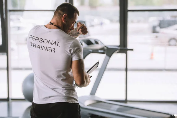 Homem com prancheta apontando na parte de trás da camiseta com lettering personal trainer no ginásio — Fotografia de Stock