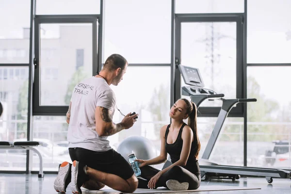 Entrenador personal masculino mostrando temporizador a la joven atlética sentada en la colchoneta de fitness con botella de agua en el gimnasio - foto de stock