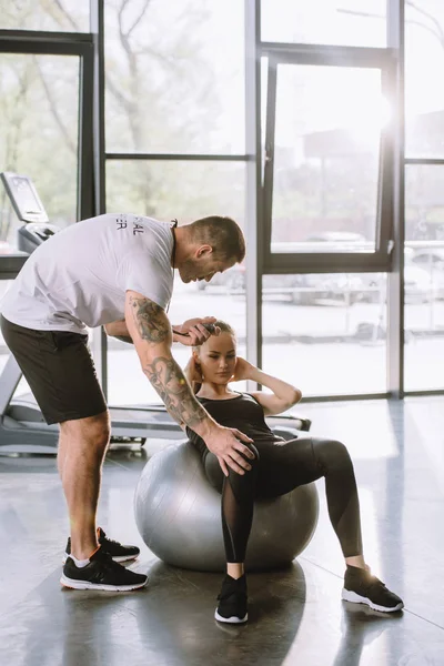 Entrenador personal masculino con temporizador y mujer atlética joven haciendo abdominales en la pelota de fitness en el gimnasio - foto de stock