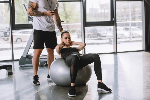 Personal trainer masculino com temporizador e jovem atlética fazendo abs na bola de fitness no ginásio — Fotografia de Stock