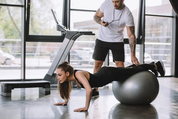 Personal trainer masculino com temporizador e jovem atlética fazendo flexões na bola de fitness no ginásio — Fotografia de Stock