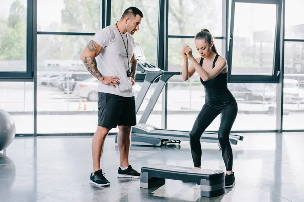 Mujer atlética joven haciendo ejercicio aeróbico paso y entrenador personal masculino en el gimnasio - foto de stock