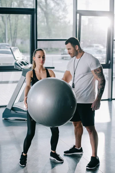 Personal trainer masculino olhando para mulher atlética exercitando-se com bola de fitness no ginásio — Fotografia de Stock