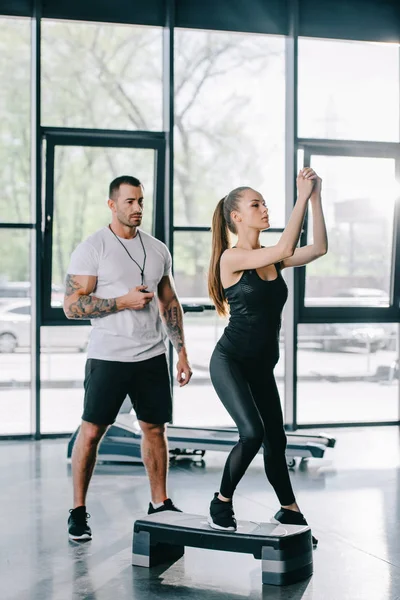 Joven deportista haciendo ejercicio aeróbico paso y entrenador personal masculino con temporizador en la mano en el gimnasio - foto de stock