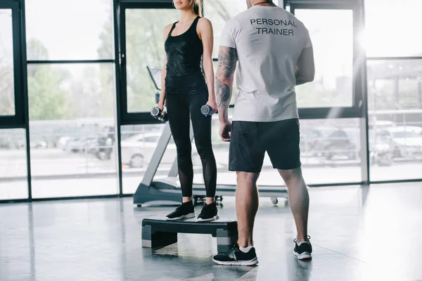 Cropped shot of male personal trainer and young sportswoman doing step aerobics exercise with dumbbells at gym — Stock Photo