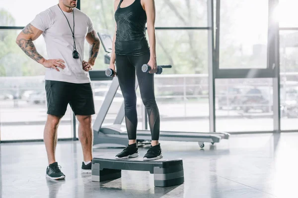Cropped image of male personal trainer and young sportswoman doing step aerobics exercise with dumbbells at gym — Stock Photo