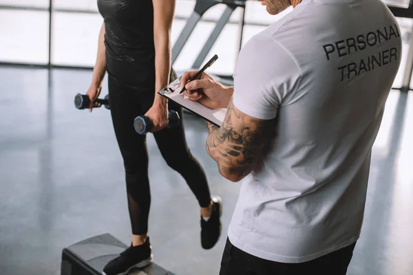 Abgeschnittenes Bild eines männlichen Personal Trainers, der in Klemmbrett schreibt und einer jungen Sportlerin, die Step-Aerobic mit Hanteln im Fitnessstudio macht — Stockfoto