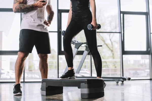 Tiro recortado de entrenador personal masculino con temporizador y joven deportista haciendo ejercicio aeróbico paso con pesas en el gimnasio - foto de stock