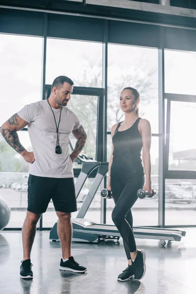 Entrenador personal masculino mirando a la deportista haciendo ejercicio con pesas en el gimnasio - foto de stock