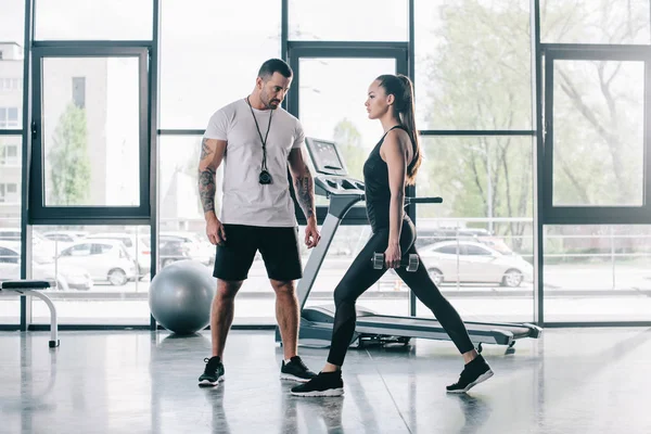 Entraîneur personnel masculin regardant la sportive faisant de l'exercice avec des haltères à la salle de gym — Photo de stock