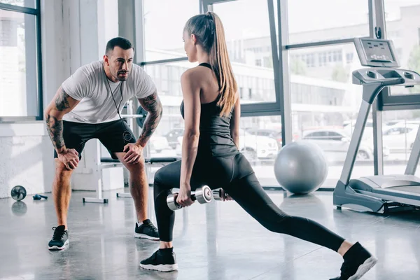 Masculino personal trainer olhando para desportista fazendo sit ups com halteres no ginásio — Fotografia de Stock