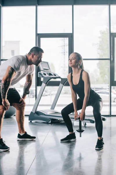 Male personal trainer looking at sportswoman doing exercises with dumbbell at gym — Stock Photo