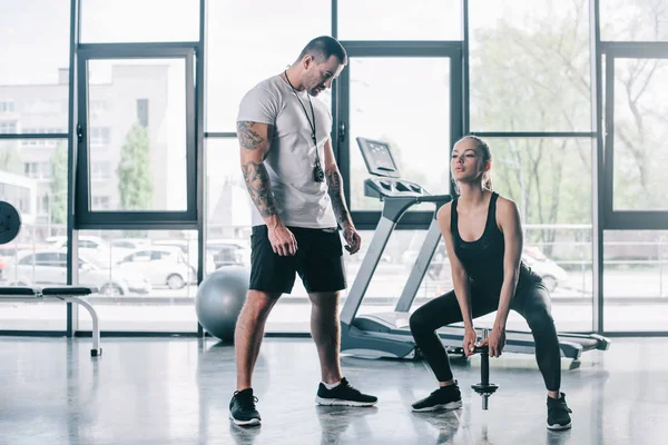 Entrenador personal masculino mirando a la deportista haciendo ejercicios con mancuerna en el gimnasio - foto de stock