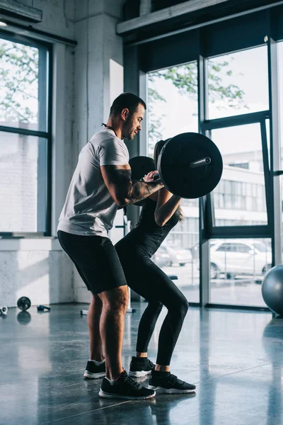 Личный тренер мужского пола помогает спортсменке делать упражнения с штангой в тренажерном зале — стоковое фото