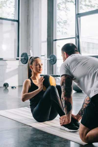 Entraîneur personnel masculin aidant la sportive à faire des abdos avec balle au gymnase — Photo de stock