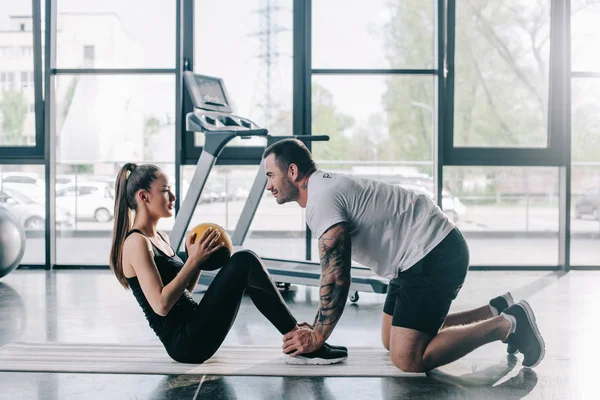 Entrenador personal masculino ayudar a la deportista a hacer abdominales con la pelota en el gimnasio - foto de stock