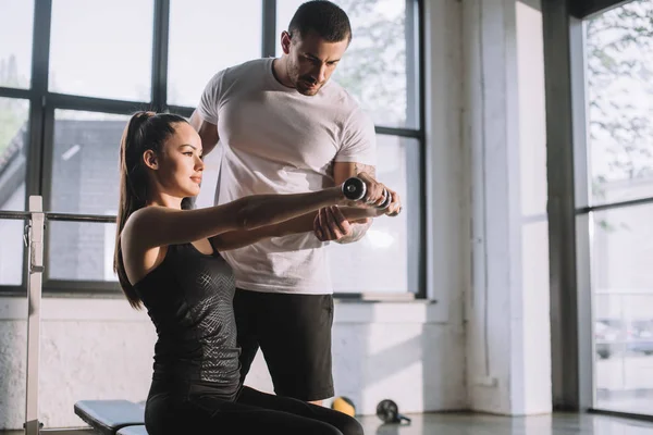 Entrenador personal masculino ayudando a la deportista a hacer ejercicios con pesas en el gimnasio - foto de stock