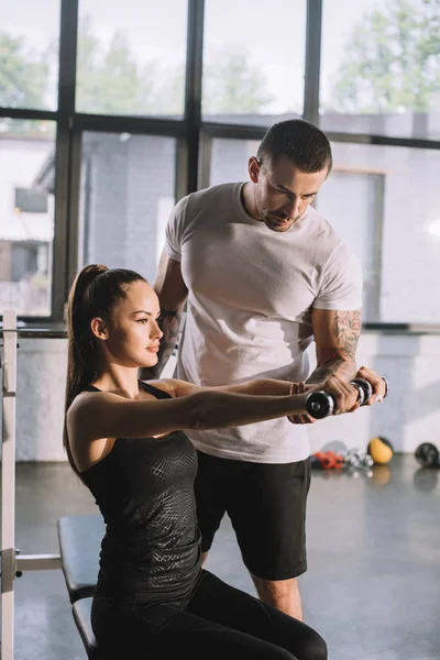 Entrenador personal masculino ayudando a la deportista a hacer ejercicios con mancuerna en el gimnasio - foto de stock