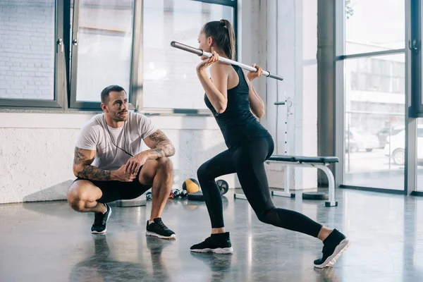 Male personal trainer looking at sportswoman doing sit ups with barbell bar at gym — Stock Photo