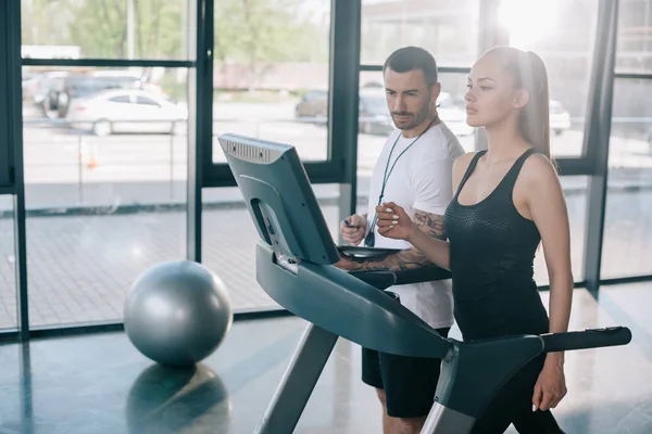 Male personal trainer looking at treadmill screen while sportswoman running at gym — Stock Photo