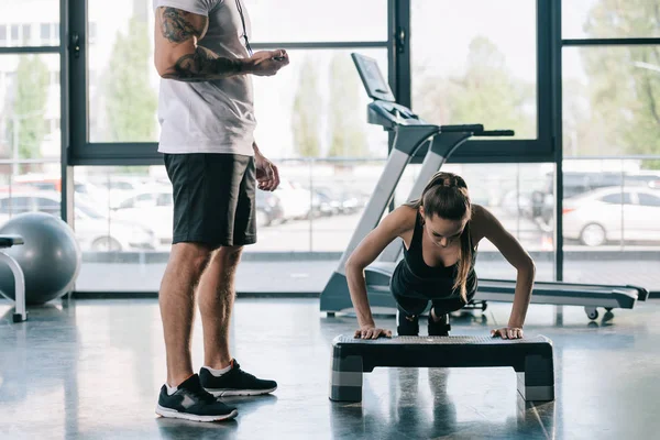 Männlicher Personal Trainer mit Zeitschaltuhr, während Sportlerin Liegestütze im Fitnessstudio macht — Stockfoto