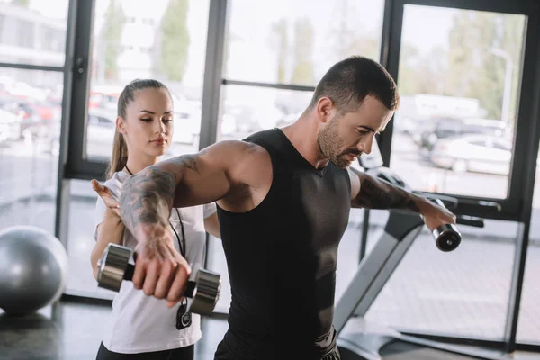 Female personal trainer helping sportsman to do exercises with dumbbells at gym — Stock Photo