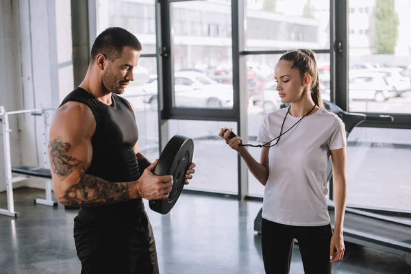 Female personal trainer using timer while sportsman doing exercises with barbell disc at gym — Stock Photo