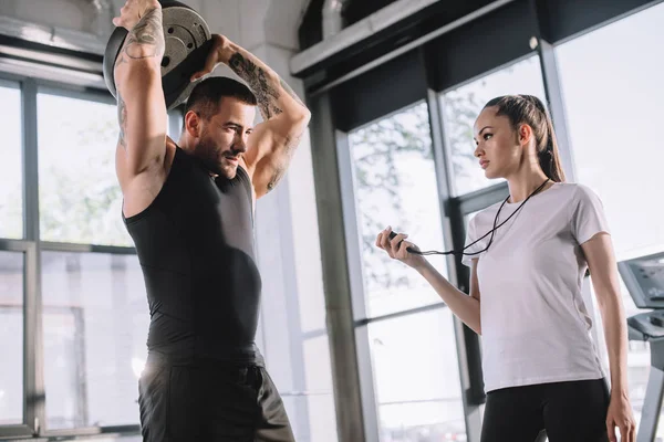 Personal trainer femminile utilizzando timer mentre lo sportivo fa esercizi con disco bilanciere in palestra — Foto stock