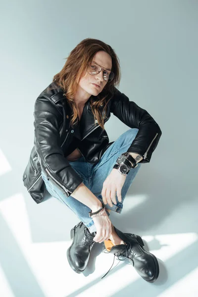 Hombre guapo y elegante con el pelo largo posando en gafas, jeans y chaqueta de cuero negro, en gris - foto de stock