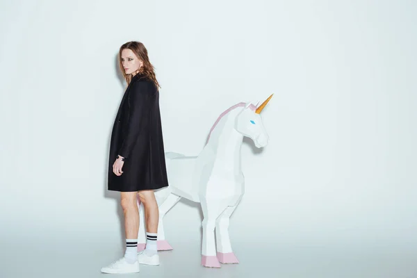 Fashionable man posing in long black jacket with big unicorn, on grey — Stock Photo