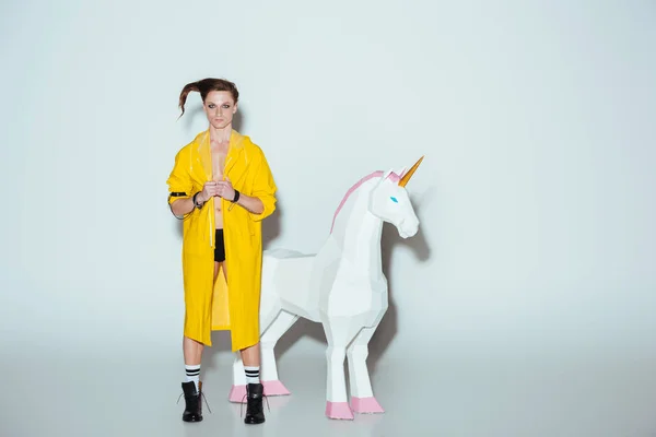 Fashionable man with hairstyle in yellow raincoat standing with big unicorn toy, on grey — Stock Photo