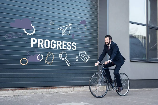 Sonriente joven hombre de negocios en traje de montar en bicicleta en la calle con inscripción de progreso e iconos de negocios en la entrada de la puerta - foto de stock