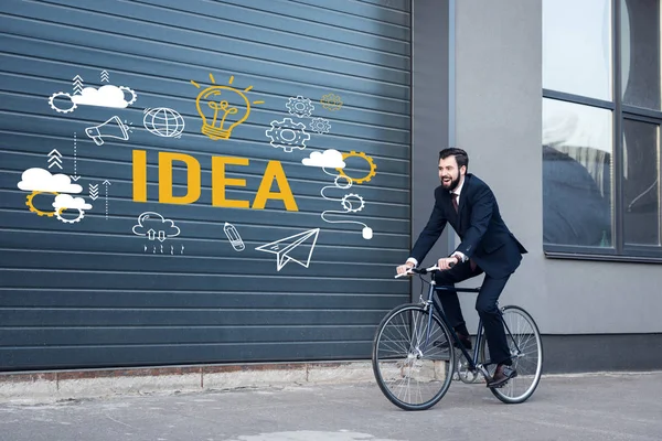 Sonriente joven hombre de negocios en traje de montar en bicicleta en la calle con idea de inscripción y los iconos de negocios en la entrada de la puerta - foto de stock