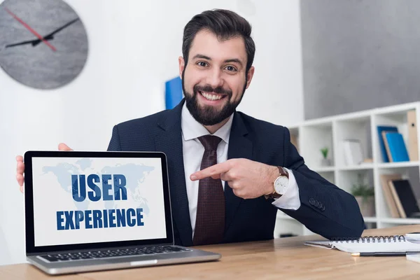 Retrato de empresário alegre apontando para laptop com inscrição experiência do usuário no local de trabalho no escritório — Fotografia de Stock