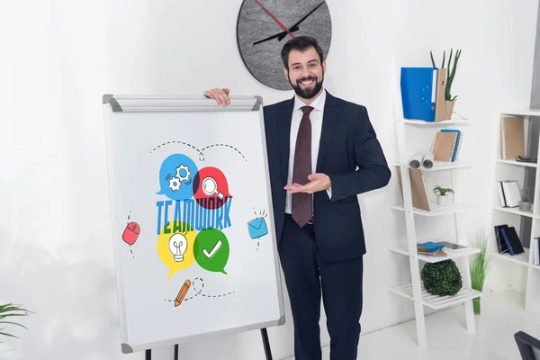 Sonriente hombre de negocios apuntando a pizarra blanca con inscripción de trabajo en equipo e iconos de negocios en la oficina - foto de stock