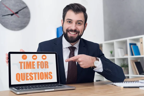 Portrait of cheerful businessman pointing at laptop with time for questions inscription at workplace in office — Stock Photo