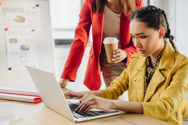 Businesswoman holding coffee cup by her colleague typing on laptop in light workspace — Stock Photo