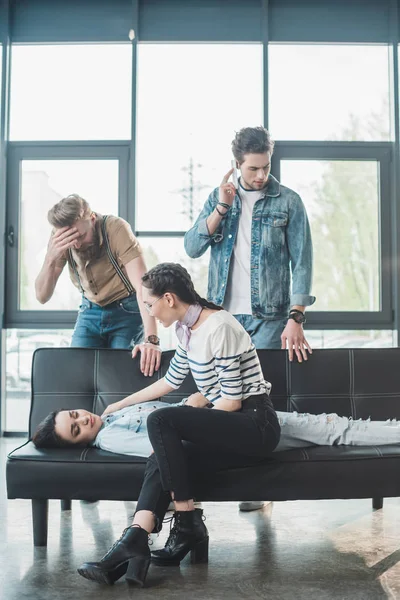 Männer und Frauen sorgen sich um junge ohnmächtige Frau, die im Büro auf Sofa liegt — Stockfoto