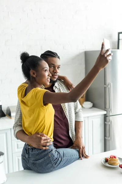 Sonriente africano americano pareja tomando selfie con smartphone en cocina - foto de stock