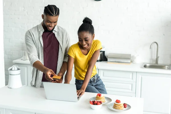 Sonriente africano americano pareja de compras en línea con tarjeta de crédito y portátil en la cocina - foto de stock