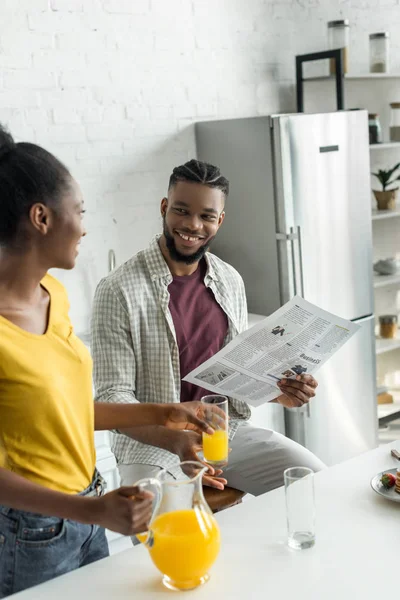 Sorrindo namorado americano africano lendo jornal e namorada derramando suco de laranja em vidro na cozinha — Fotografia de Stock