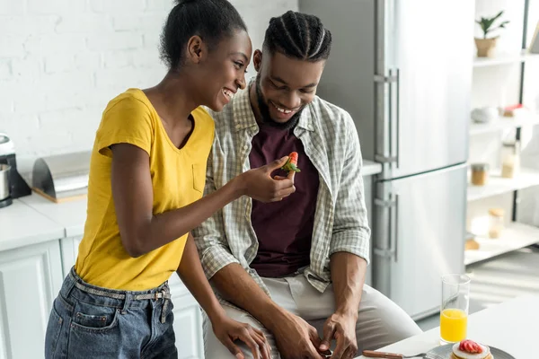 African american girlfriend feeding boyfriend with strawberry at kitchen — Stock Photo