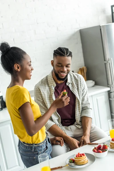 Smiling african american girlfriend feeding boyfriend with strawberry at kitchen — Stock Photo