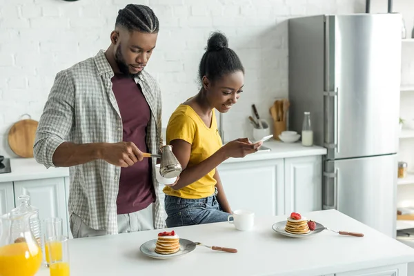 African american girlfriend taking photo of pancakes with smartphone at kitchen — Stock Photo