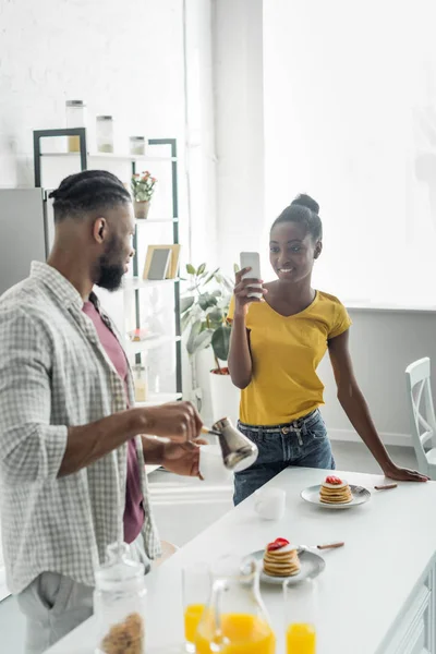 African american girlfriend taking photo of boyfriend with coffee with smartphone at kitchen — Stock Photo