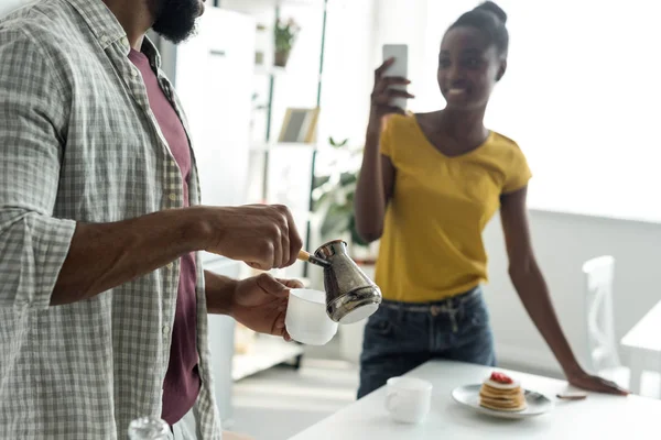 Обрезанное изображение африканской американской подружки, фотографирующей парня со смартфоном на кухне — стоковое фото