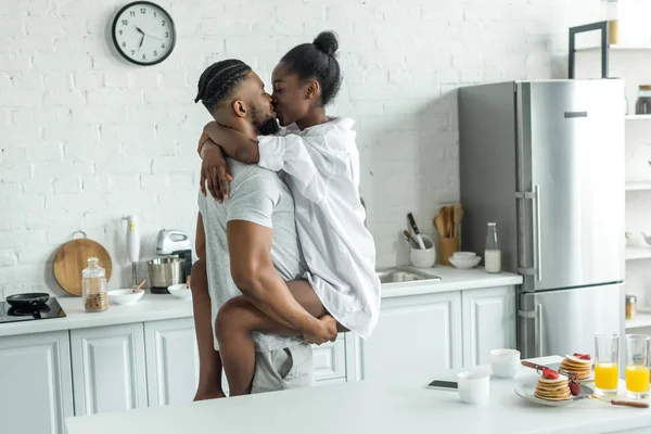 Vista lateral do namorado afro-americano segurando namorada e eles se beijando na cozinha — Fotografia de Stock
