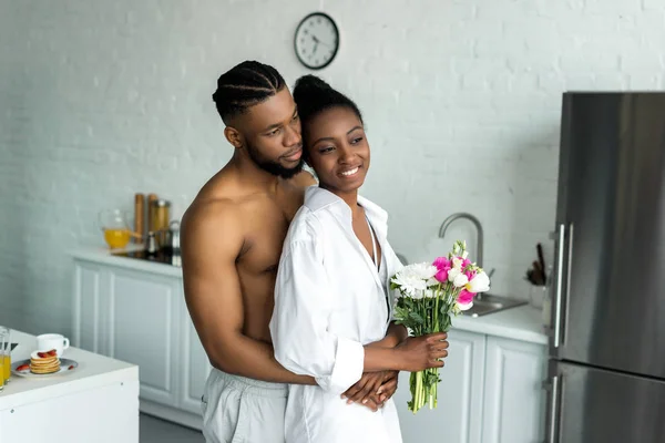 Africano americano novio abrazando novia y ellos mirando lejos en cocina - foto de stock