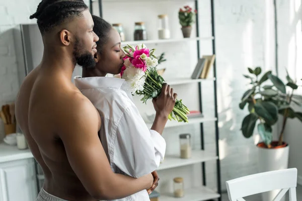 Africano americano fidanzato abbracciare fidanzata e lei annusare mazzo di fiori a cucina — Foto stock