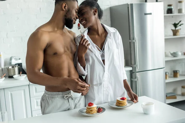 Afro americana namorada segurando garfo com morango entre ela e namorado na cozinha — Fotografia de Stock